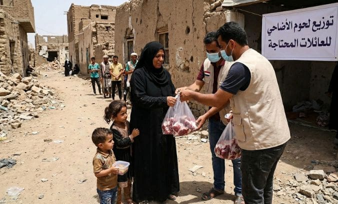 Qurbani in War Zones: How Your Qurbani Meat Donation Reaches Families in Palestine and Yemen 3 Two aid workers wearing face masks and vests distribute bags of Qurbani meat to a woman in a black abaya standing with two young children. They are on a dirt street in a conflict-affected area with damaged buildings and rubble. In the background, other people are waiting in line, and a banner with Arabic text is mounted on a nearby wall.