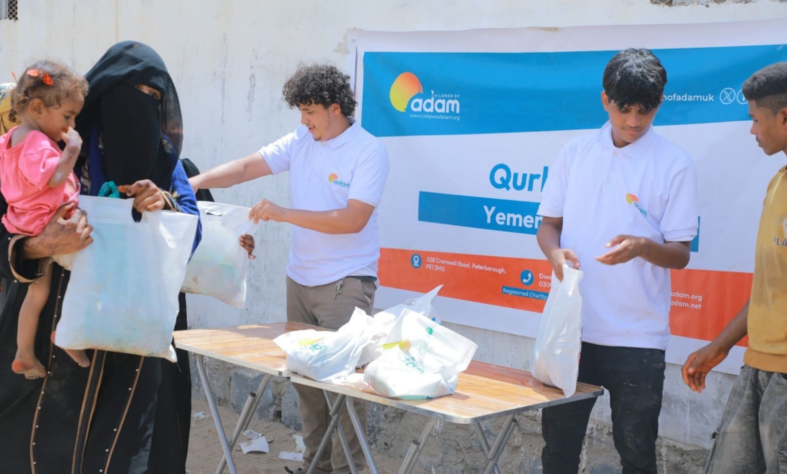 Inside Yemen Qurbani 2026 Distribution: How Your Donation Reached Families in Crisis 6 Two volunteers in white "Children of Adam" t-shirts handing out donation bags from a folding table to a woman wearing a niqab and holding a young child during a Qurbani event in Yemen.