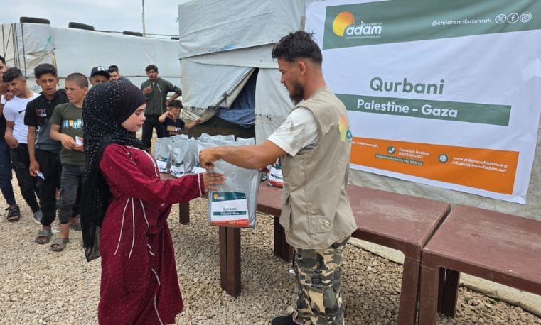 Qurbani 2026: Delivering Hope and Essential Food Support to Families in Gaza 5 A volunteer handing a Qurbani aid package to a young girl in a red dress during a Children of Adam distribution event in Palestine-Gaza, with other children waiting in line behind her.