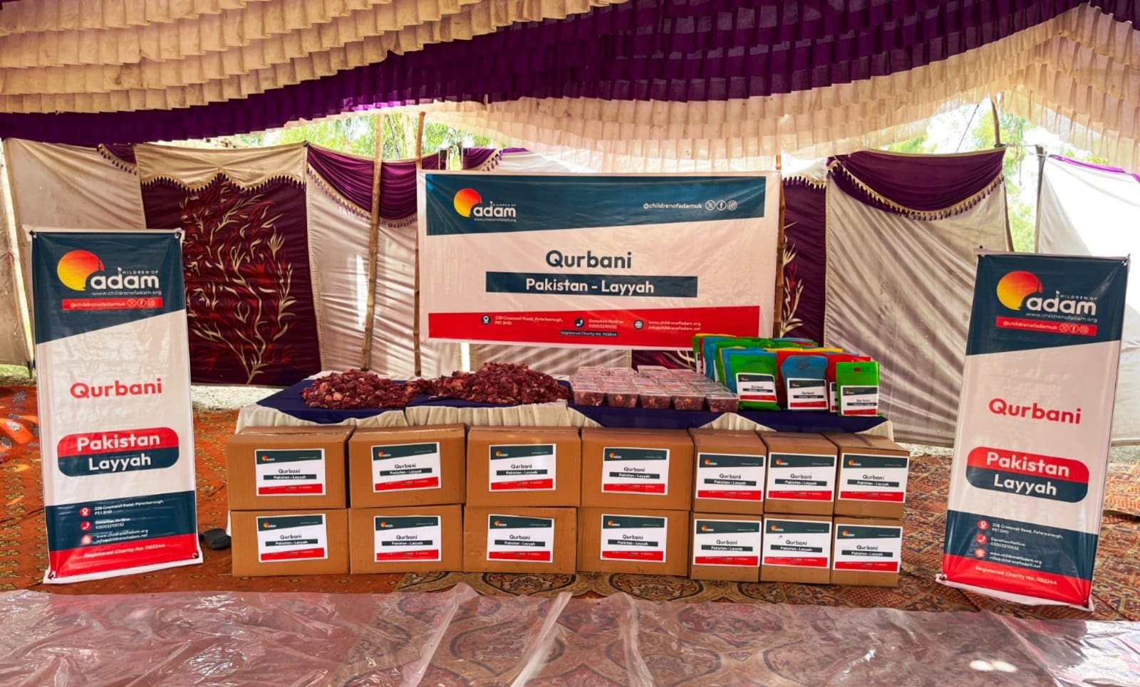 Qurbani 2026: Fresh Meat Distributed to Families in, Pakistan 11 A distribution setup under a tent for a "Children of Adam" Qurbani event in Layyah, Pakistan, featuring organized rows of cardboard boxes, meat portions, and colorful donation bags arranged on tables with event banners.