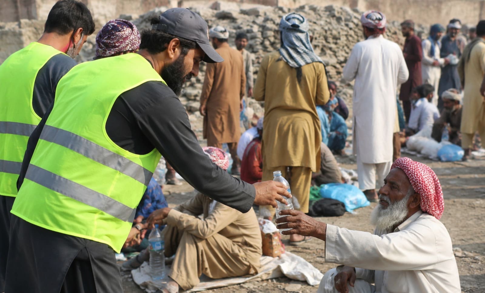 The Virtues of Giving Water in Islam: Hadith and Quranic Teachings 4 a volunteer wearing a bright neon safety vest hands a bottle of water to an elderly man sitting on the ground at a crowded outdoor relief distribution site.
