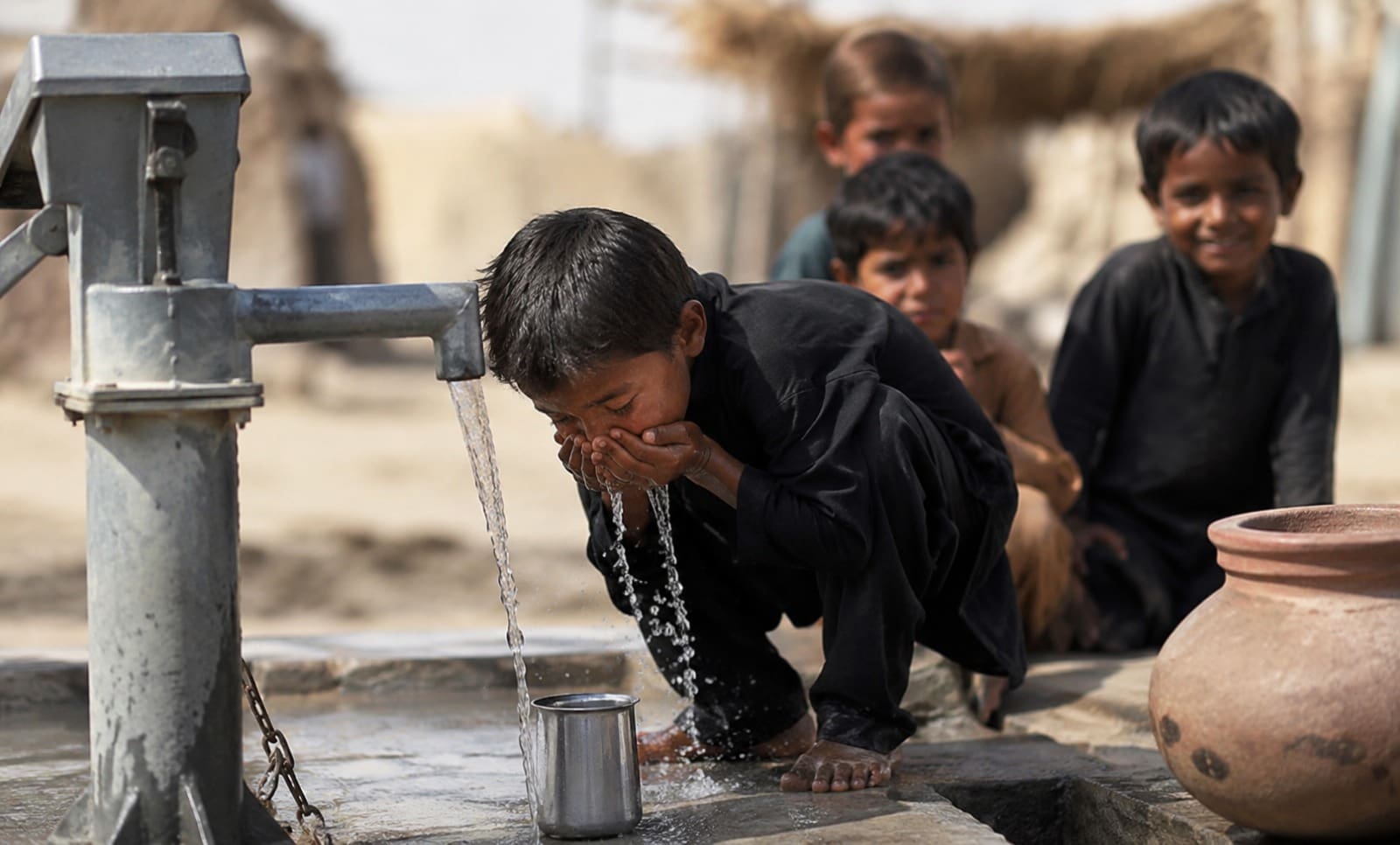 Why Water Pump Donation is the Best Muharram Charity in Islam 4 A young boy in dark clothing kneels on a concrete platform to drink fresh water flowing from a manual hand pump. He cups his hands under the stream, with water splashing down. In the background, three other children look on attentively near a clay water pot, set against a blurred, rural outdoor environment.
