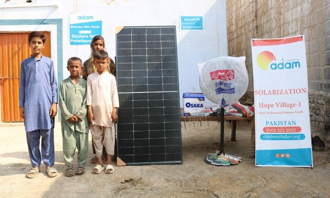Donate Solar Fans for Families in Pakistan 5 four children stand outside their home in khairpur mirs, sindh, next to a newly delivered solar panel, an osaka battery box, and a standing fan.