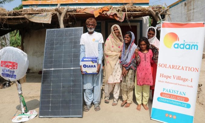 How Power Cuts Make Heatwaves More Dangerous in Pakistan 3 a smiling family poses outside their home in khairpur muhammad somoro, sindh, next to a solar panel, an osaka battery box, and a standing fan.