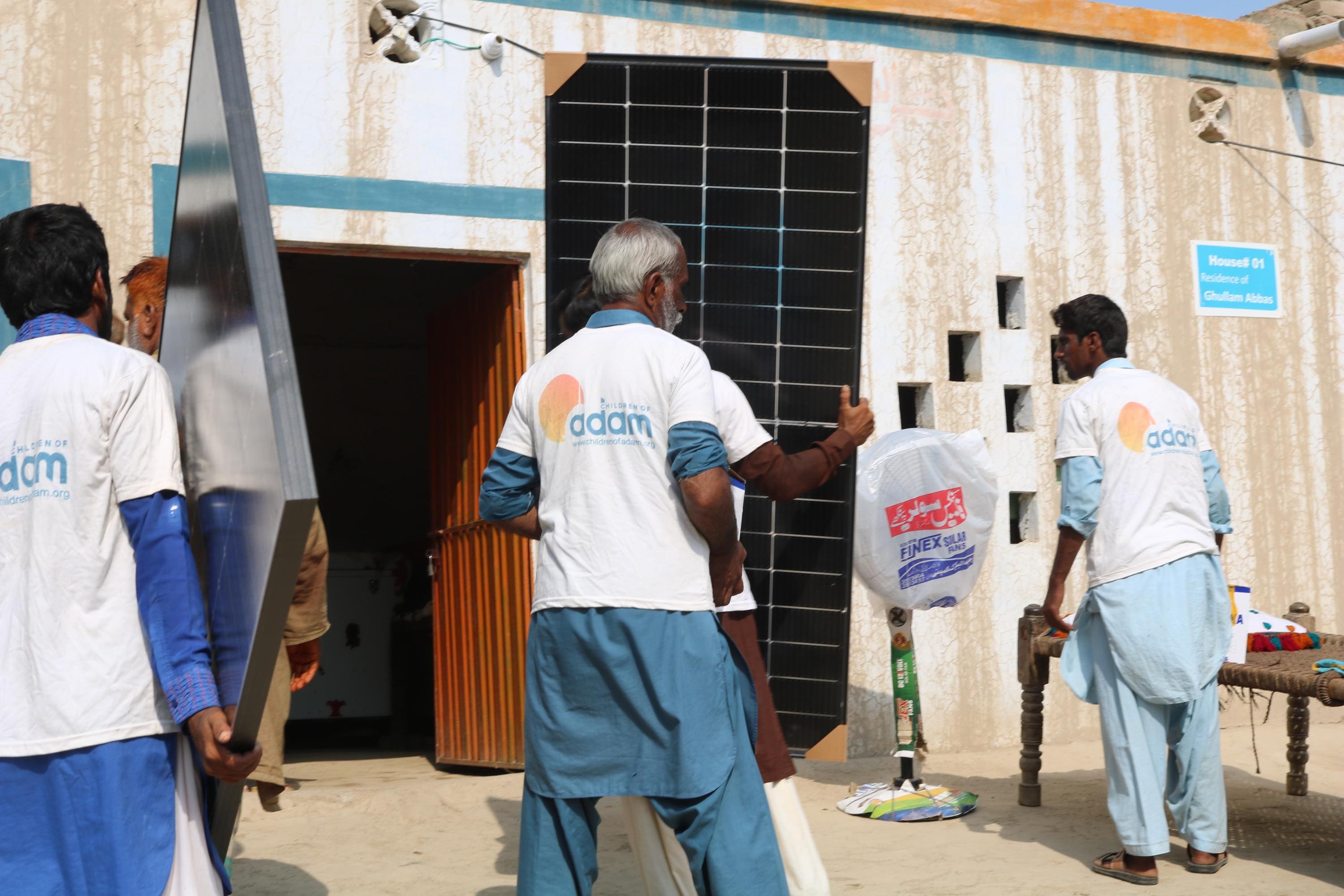 Why Solar Panels Are Sadaqah Jariyah 8 men wearing "children of adam" t-shirts carry and set up solar panels outside a house labeled "residence of ghullam abbas."